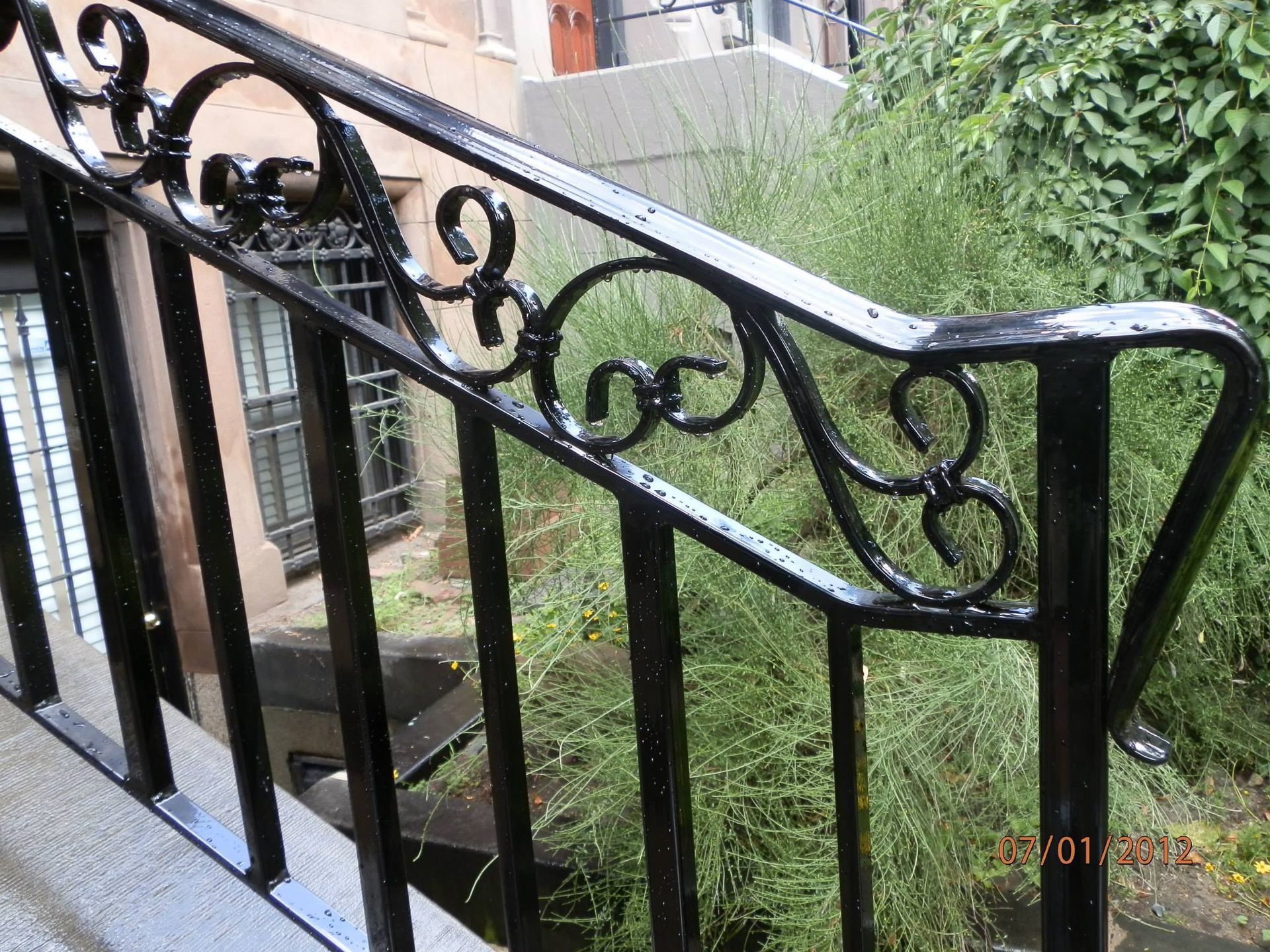 Black wrought-iron railing with curled scrollwork beside a brick building and greenery