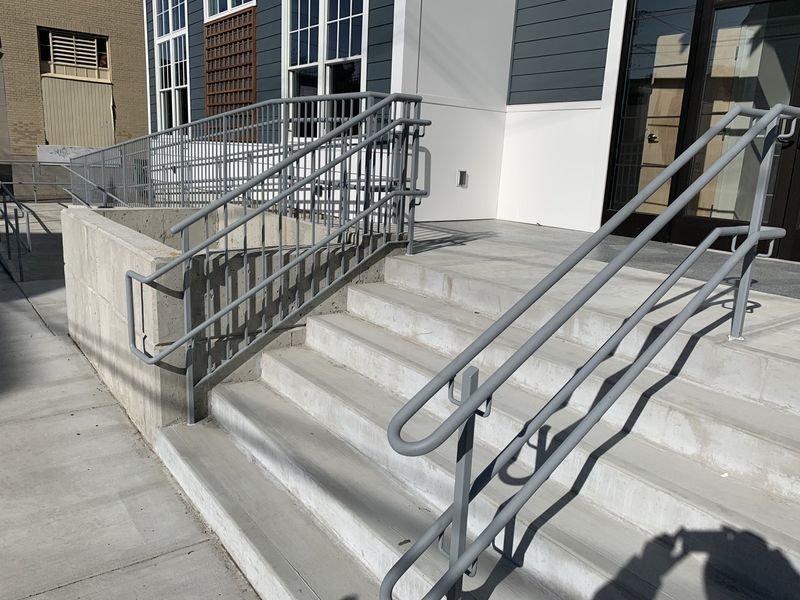 Concrete steps leading to a building entrance with grey metal handrails on both sides and a side concrete retaining wall.