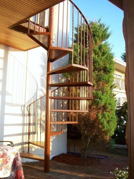 A brown metal spiral staircase leading to a wooden deck, situated outdoors next to a building and a tall evergreen tree.