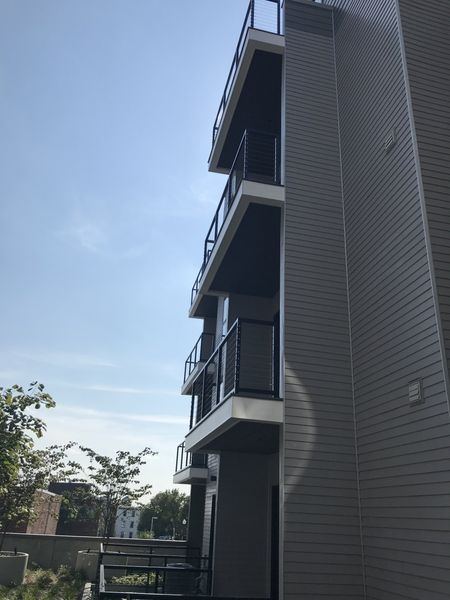 A side view of a modern, multi-story building featuring stacked balconies with black railings against a clear blue sky.