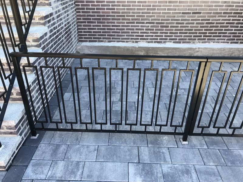 Black metal railing with a rectangular grid pattern on a brick-paved patio in front of a brick wall.