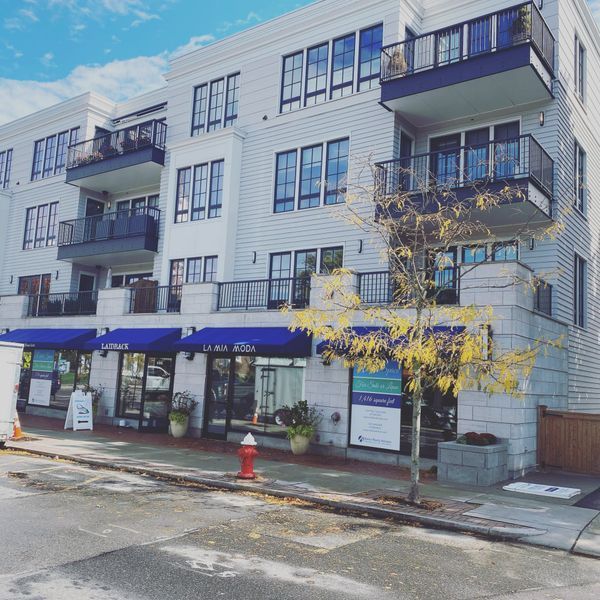 A multi-story building with light-colored siding, black balconies, and blue awnings over street-level storefronts.