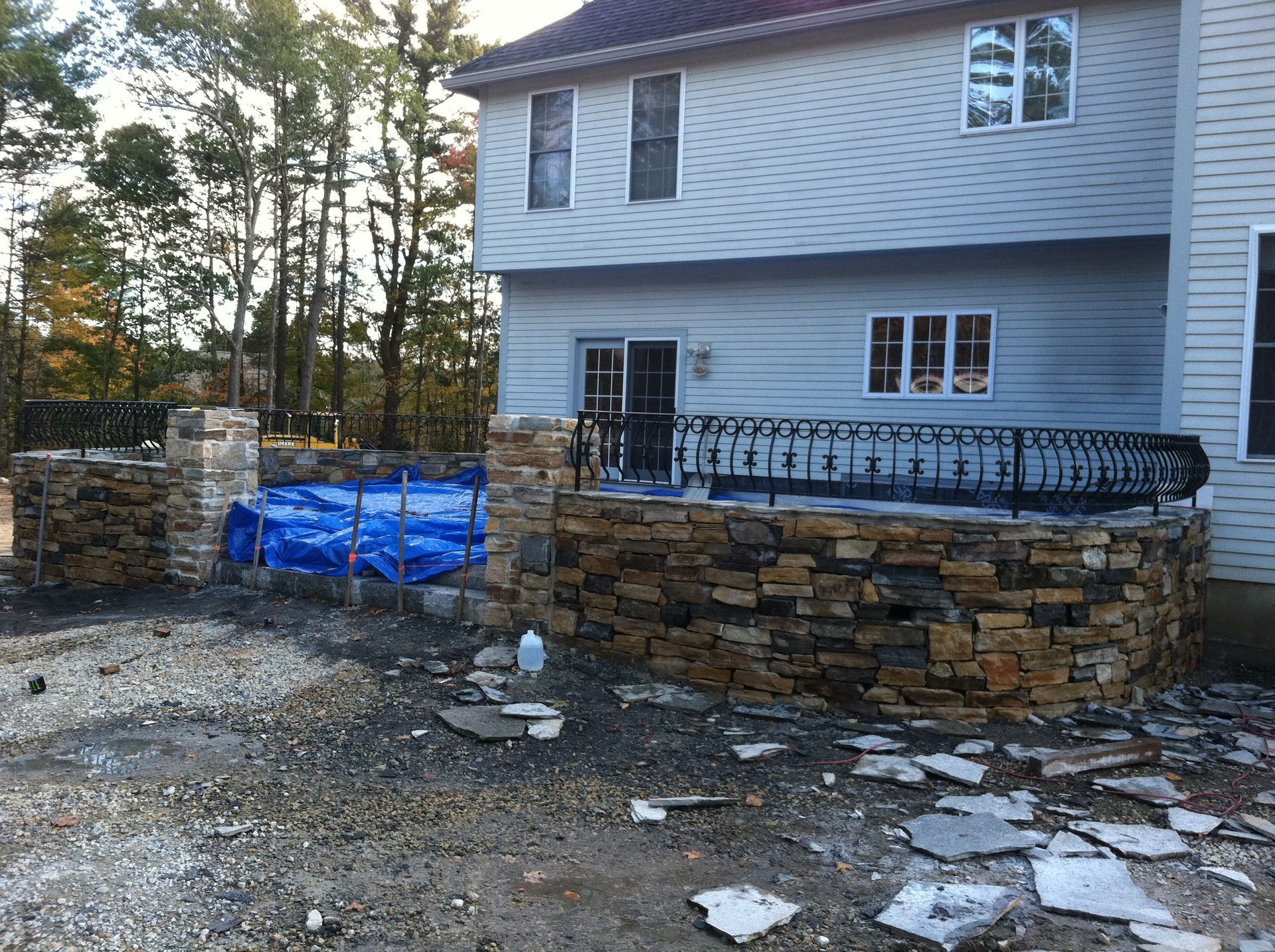 Stone retaining wall and blue tarp in a driveway beside a gray house with trees in the background