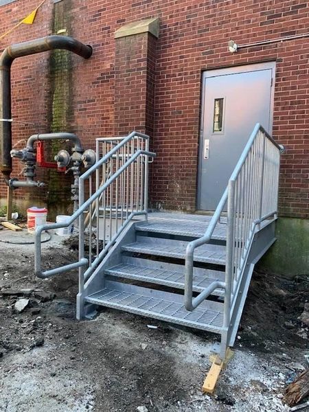 A gray metal staircase with handrails leading to an exterior building door against a red brick wall.