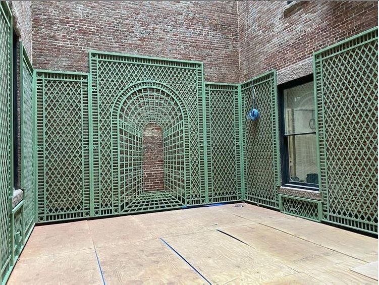A courtyard with sage green wooden lattice wall panels, including a centered arched design against a brick wall.