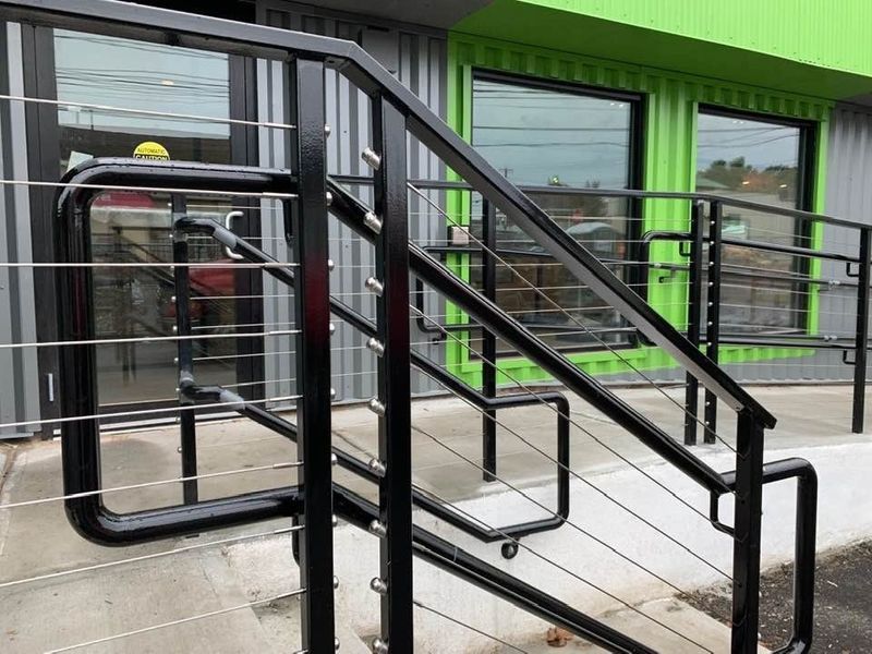 Black metal handrails with horizontal cable railing along a ramp and stairs outside a building with a green facade.