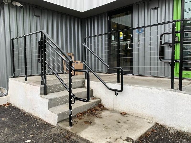 Concrete entrance steps with black metal railings and wire infill panels leading to a glass door on a grey industrial building.