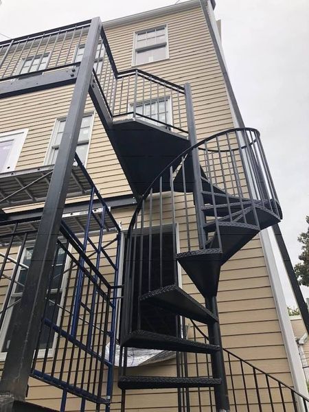 Black metal spiral staircase attached to the exterior side of a tan-sided building with windows.