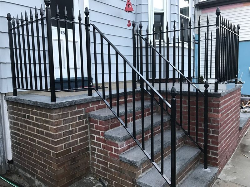 Black metal railings with spear-top finials line brick steps and a small landing outside a house with blue siding.