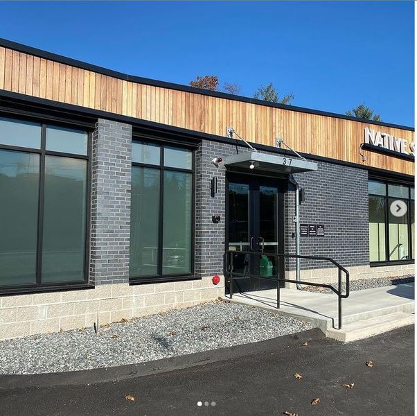 Exterior of a modern brick and wood building entrance with glass doors, black window frames, and a concrete ramp.