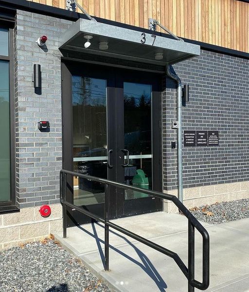 A modern building entrance with a black double door, dark brick exterior, silver canopy, and a black metal handrail.