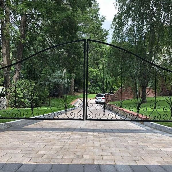 A black wrought-iron gate stands closed across a stone-paved driveway, with green trees and a white car in the background.