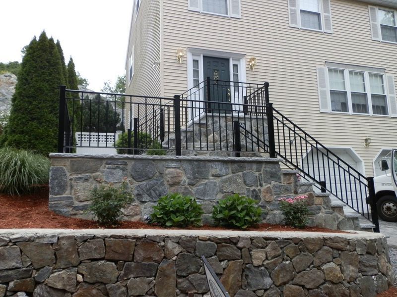 A tan home with a black front door, metal railing, and two levels of stone retaining walls with small green bushes.