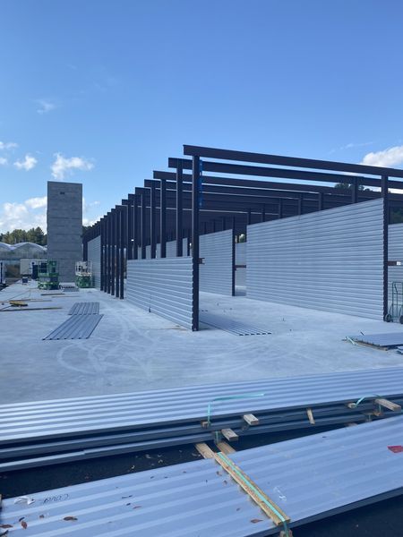 A construction site showing a steel-framed warehouse skeleton with corrugated metal wall panels under a clear blue sky.