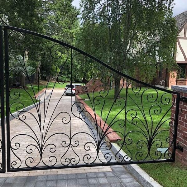 A black wrought iron driveway gate with decorative scrollwork, set against a brick pillar and a driveway.