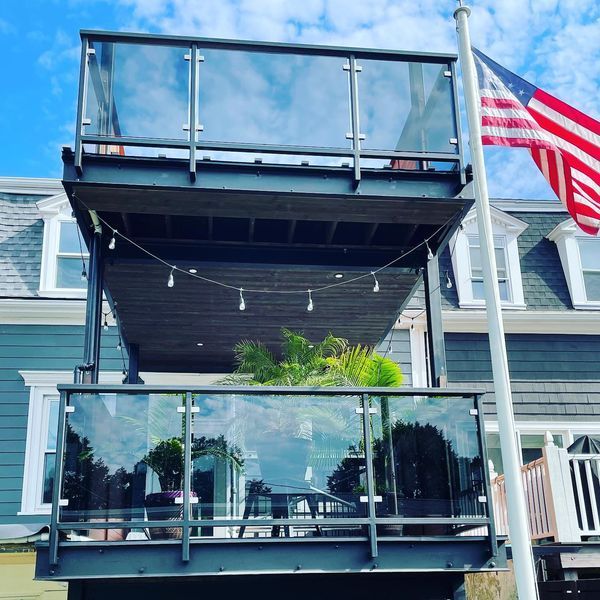 A two-story deck with glass railings and a string of lights, attached to a blue house with an American flag nearby.