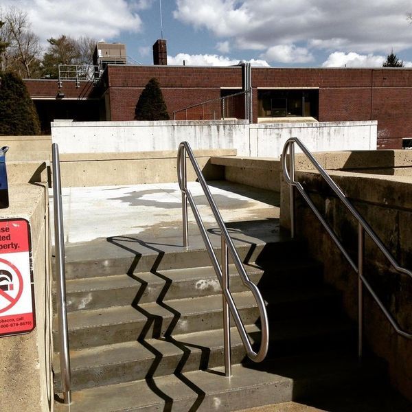 Concrete stairs with metal handrails leading to an outdoor brick building under a sunny, partly cloudy sky.