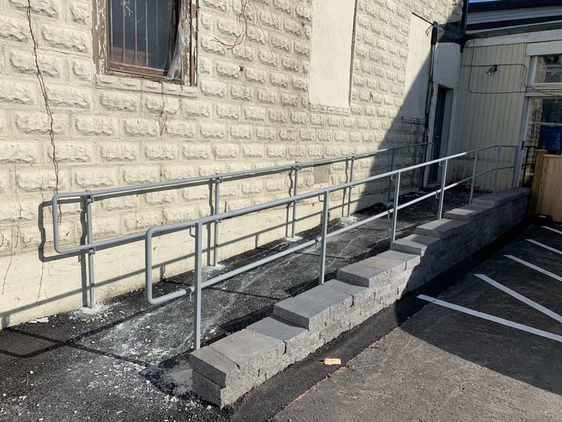 A metal handrail runs along a gray concrete block retaining wall and ramp outside a building with a light stone exterior.