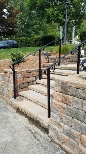 Stone steps leading up a grassy hill, framed by a tan stone retaining wall and black wrought iron handrails.