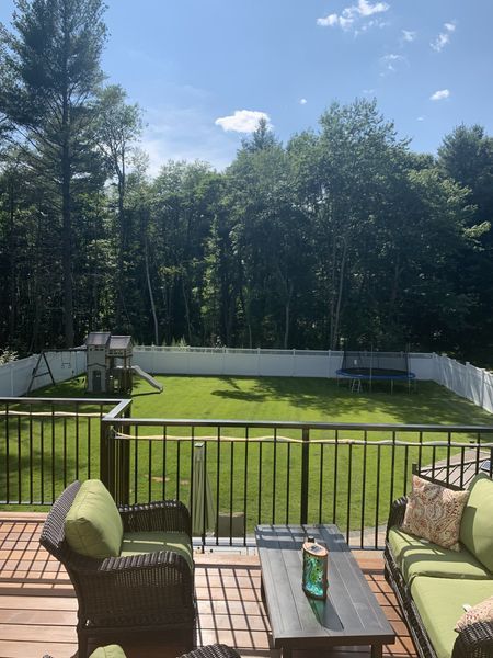 A sunlit backyard viewed from a wooden deck with outdoor seating, a white fence, a playset, and a trampoline.