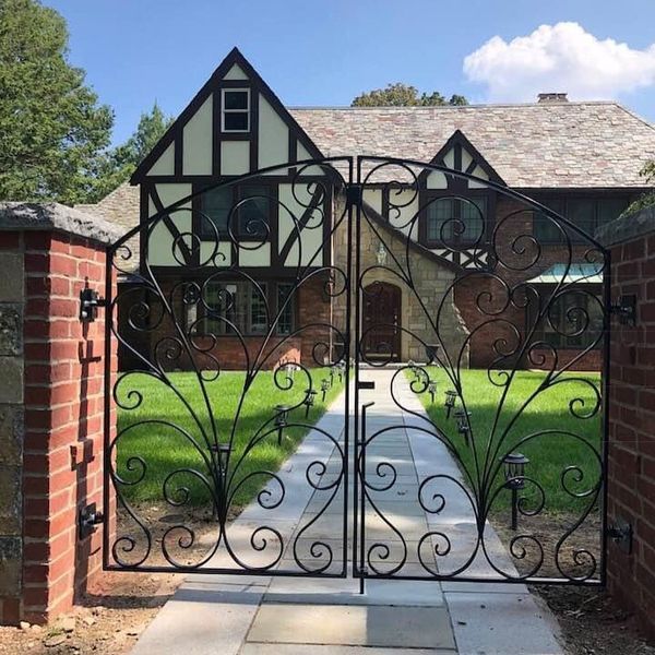 A black ornate wrought-iron gate stands before a Tudor-style brick home with a stone path leading to the front door.