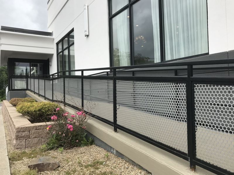A concrete accessibility ramp featuring black railings with perforated metal panels along the side of a white building.