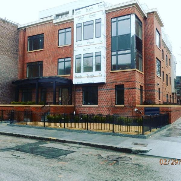 A modern, multi-story brick apartment building on a corner lot with a black metal fence and an unfinished window section.