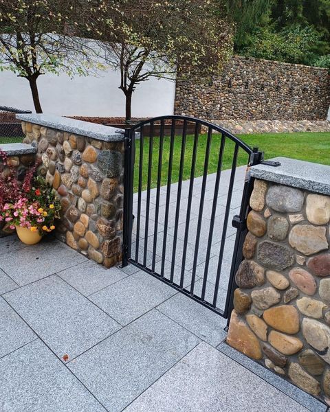 A black metal gate arched at the top sits between two stone walls on a stone patio, with a green lawn in the background.