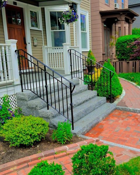 Stone steps with black metal handrails leading to the front porch of a tan house with a brick walkway in the foreground.