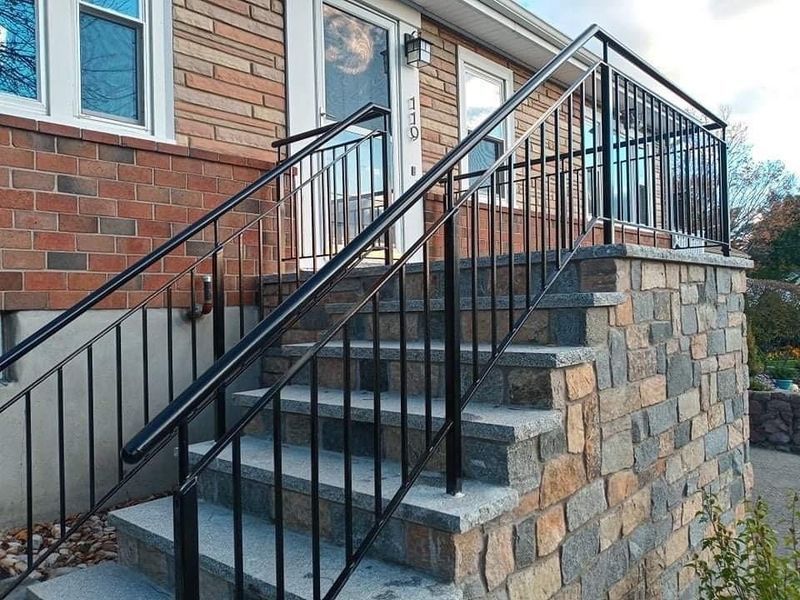 Stone steps lead to a house entrance, featuring black metal railings on both sides against a brick and stone facade.