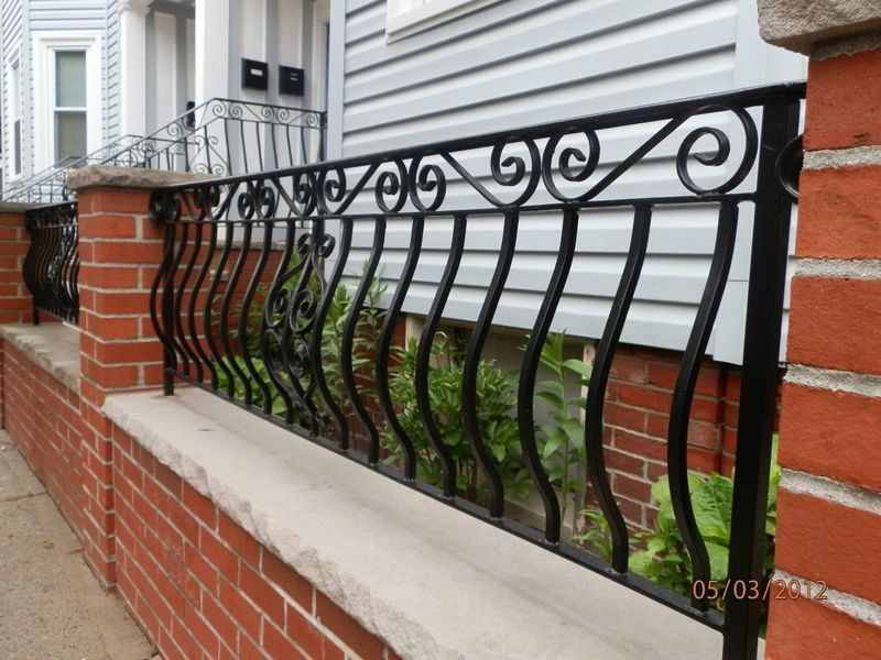 A black wrought iron fence with decorative scrolls sits atop a red brick wall in front of a house with grey siding.
