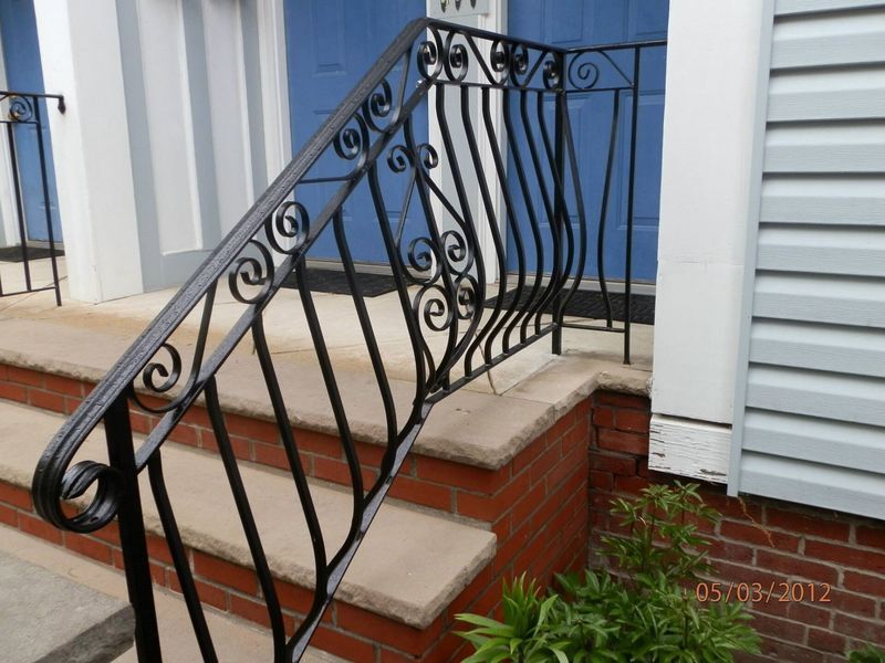Black metal wrought-iron railing with scrollwork designs leading up brick steps to a building entrance.