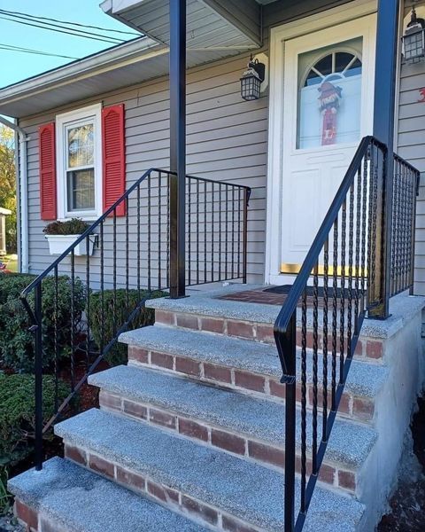 A set of stone steps leading to a house entrance with black metal railings on both sides and red window shutters.