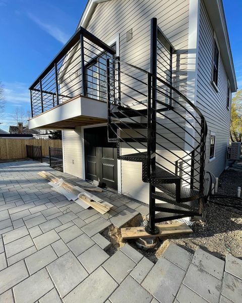 A black metal spiral staircase leads to an upper deck on the side of a light-colored house with a paved patio area below.