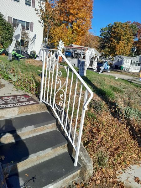 A white, decorative metal handrail runs alongside concrete front steps on a sunny day with fall foliage in the background.