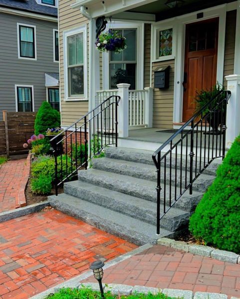 Concrete steps with black iron railings lead up to a porch with a wooden door and tan siding, set next to a brick path.