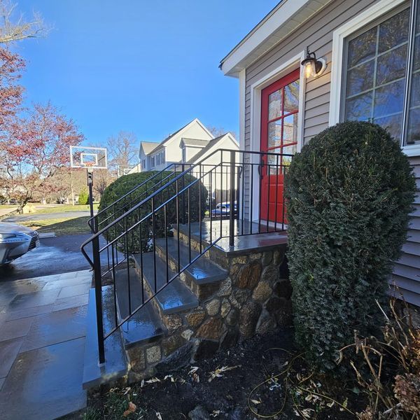 Stone steps lead to a red front door of a gray house, featuring a dark metal railing and landscaped evergreen bushes.