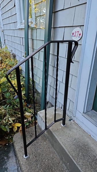 A black metal handrail mounted to concrete steps outside a house with gray shingles and a house number sign reading 27.