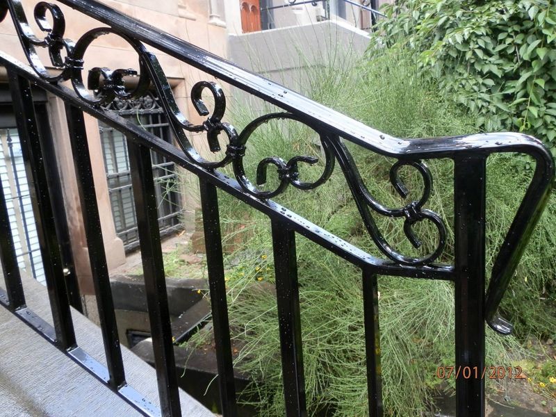 Black wrought iron stair railing featuring decorative scrollwork, set against a background of greenery and a building.