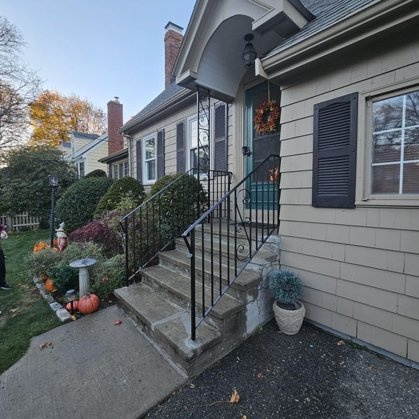 A beige house with dark shutters, stone steps, a black metal railing, and fall decorations including pumpkins and wreaths.