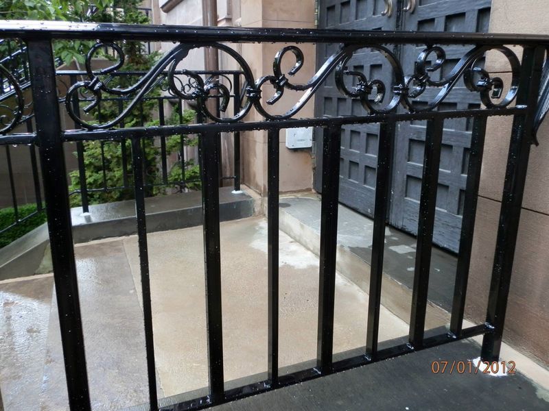 Black metal railing with decorative scrollwork at the top, standing in front of a building entrance with steps.