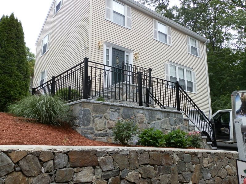A two-story, light-colored house with a black metal railing on a stone-faced raised porch and a stone retaining wall.