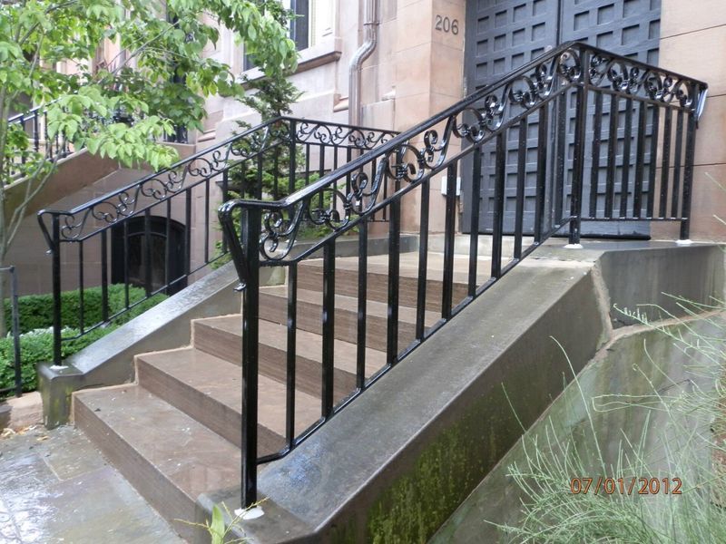 Concrete steps leading up to an entrance door with black metal railings on both sides, surrounded by trees and greenery.