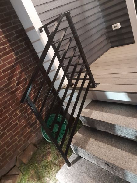 A black metal railing installed on the side of outdoor concrete steps leading to a wooden porch.