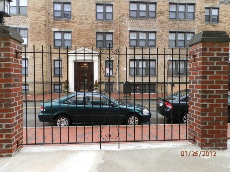 A black iron fence stands before a brick apartment building and two parked cars on a city street.