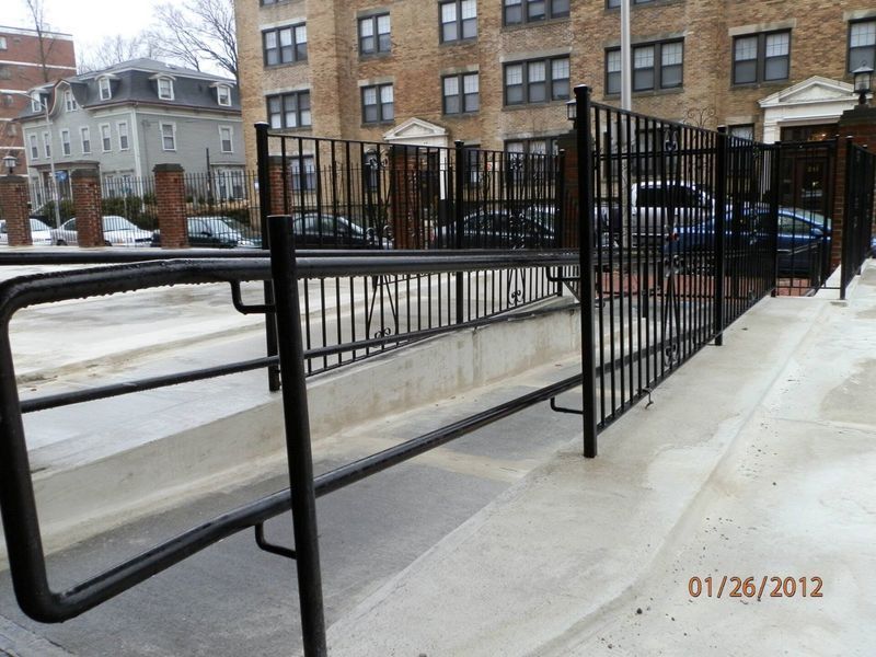 A black metal railing lines a concrete wheelchair ramp leading to an apartment building entrance on a cloudy day.