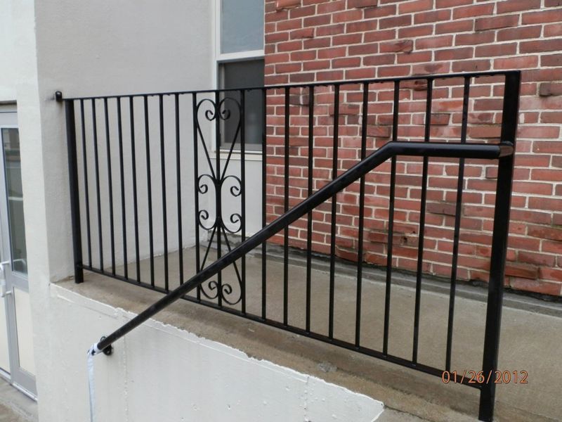 Black metal railing featuring a central decorative scroll design, installed on a concrete porch against a brick wall.