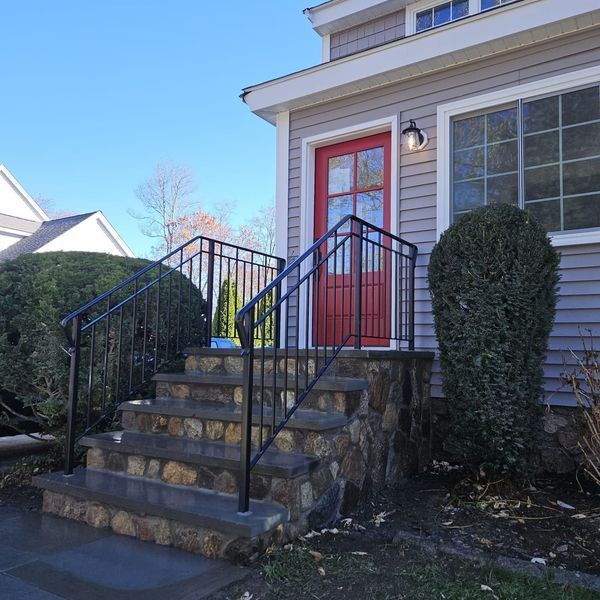 A stone-faced staircase with a black metal railing leading to a house with gray siding and a bright red front door.