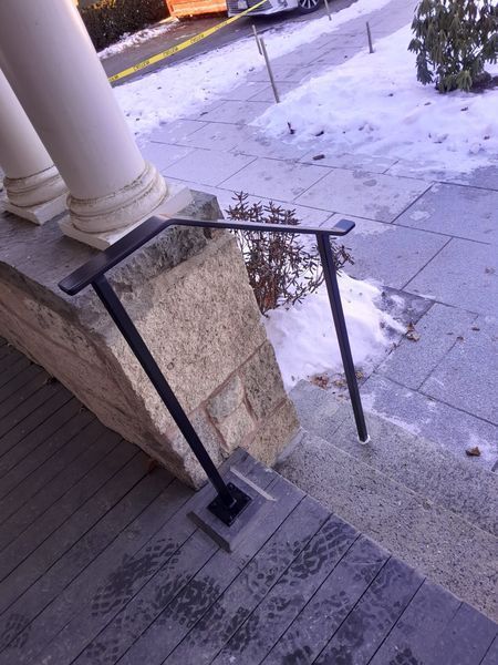A black metal handrail installed on a stone porch staircase, with snow visible on the ground in the background.