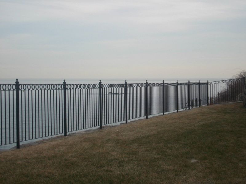 A black metal fence runs along a grassy bluff overlooking a calm, overcast body of water.